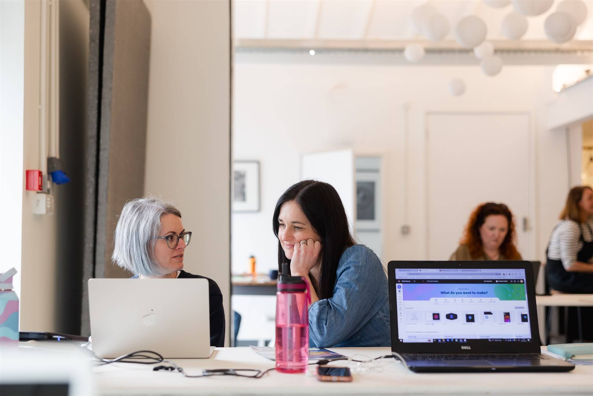 two woman sat together at long table and working at the laptop looking relaxed in each others company